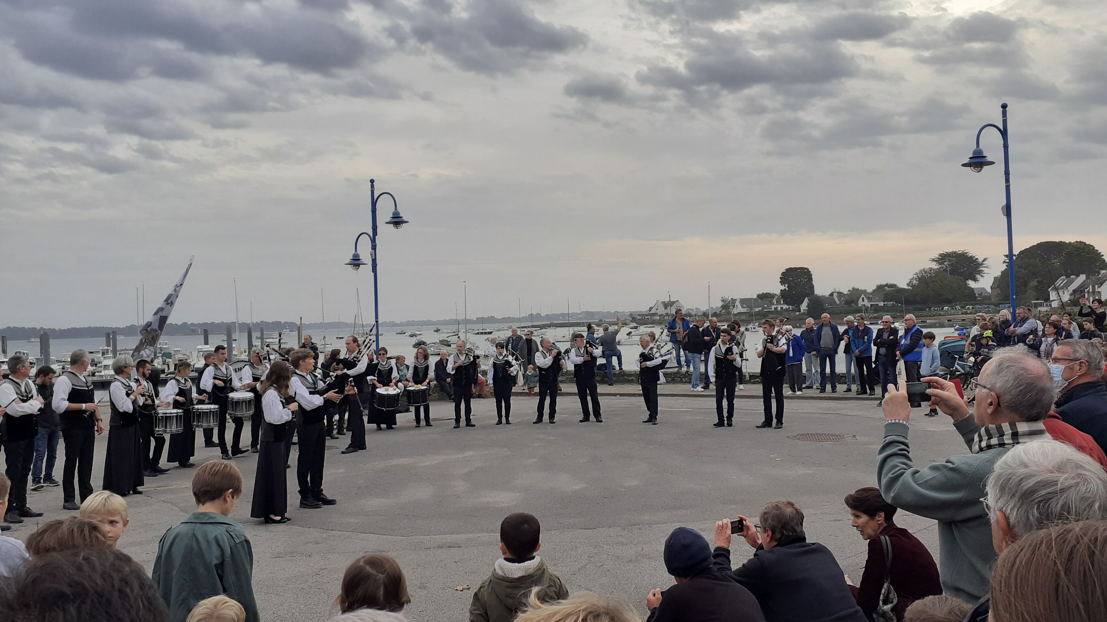 A group of people gathered outdoors, some standing and some sitting, with a body of water and boats in the background.
