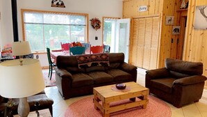 Un salon chaleureux avec un canapé marron, une table basse en bois et un tapis rouge. On y trouve également un coin repas avec une table et des chaises, ainsi qu’une grande fenêtre qui laisse entrer la lumière naturelle.