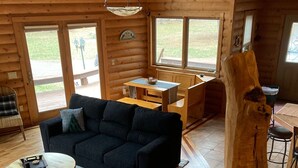 Un interior de cabaña acogedor, con suelo de madera, un sofá negro, una mesa de centro redonda y un comedor con banco y mesa.
