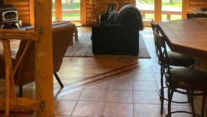 Interior acogedor de una cabaña con detalles de madera, chimenea, sofá y ventanas amplias que ofrecen vistas al exterior.