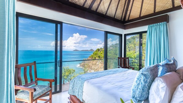 Un dormitorio con un ventanal que ofrece vistas al mar, una silla de madera y una cama con almohadones estampados.