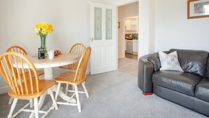 A dining area with a round table, four wooden chairs, a vase of yellow flowers, and a coffee pot. A gray leather sofa is against the wall, and a kitchen is visible through an open door. - Greenfields - UK43269 (Upottery, near Honiton)