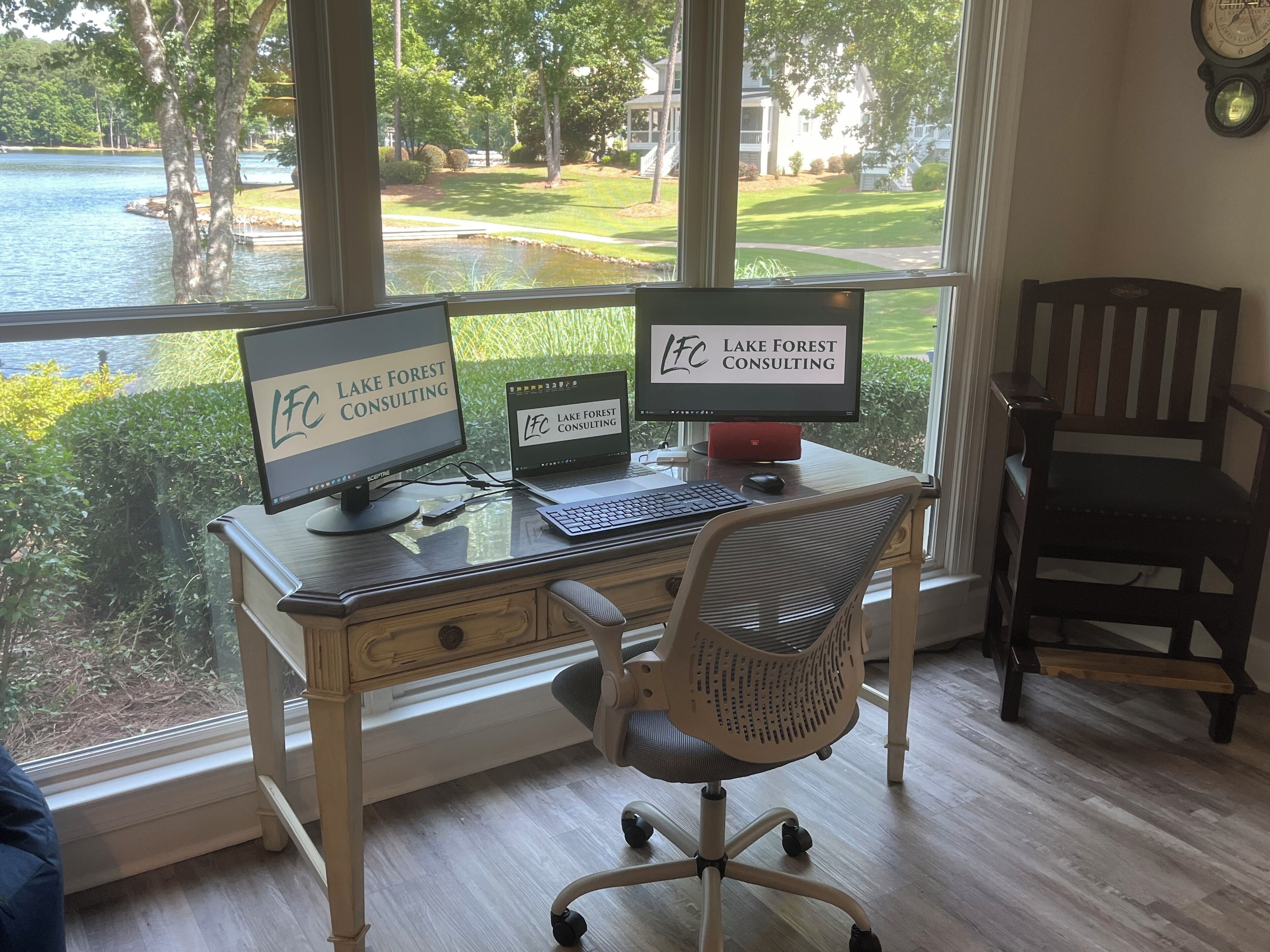 Un bureau avec deux écrans, un clavier et une chaise, placé devant une fenêtre donnant sur un lac et une pelouse.