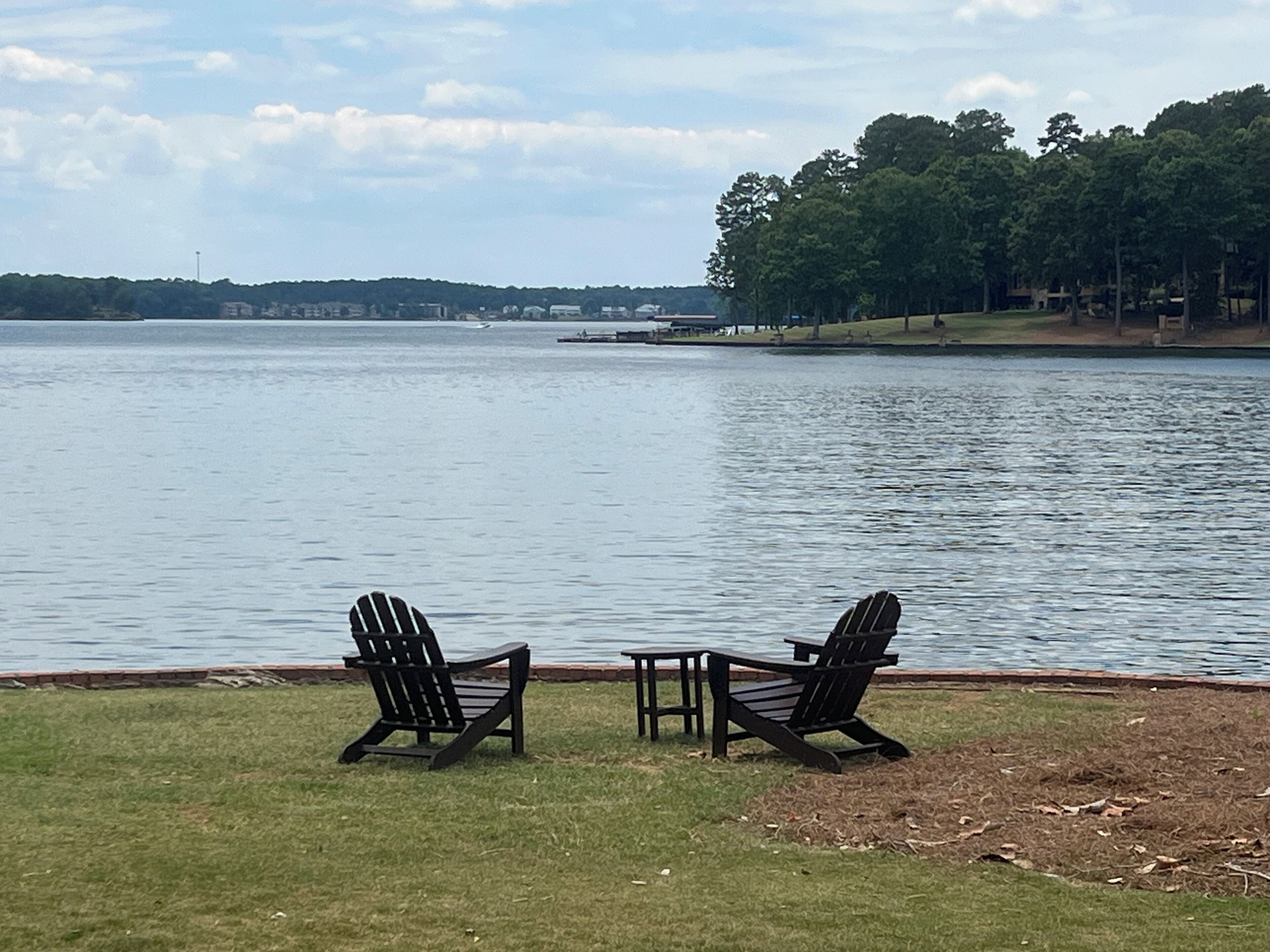 Deux fauteuils Adirondack sont placés face à un lac, avec une zone herbeuse au premier plan et une rive bordée d’arbres en arrière-plan.