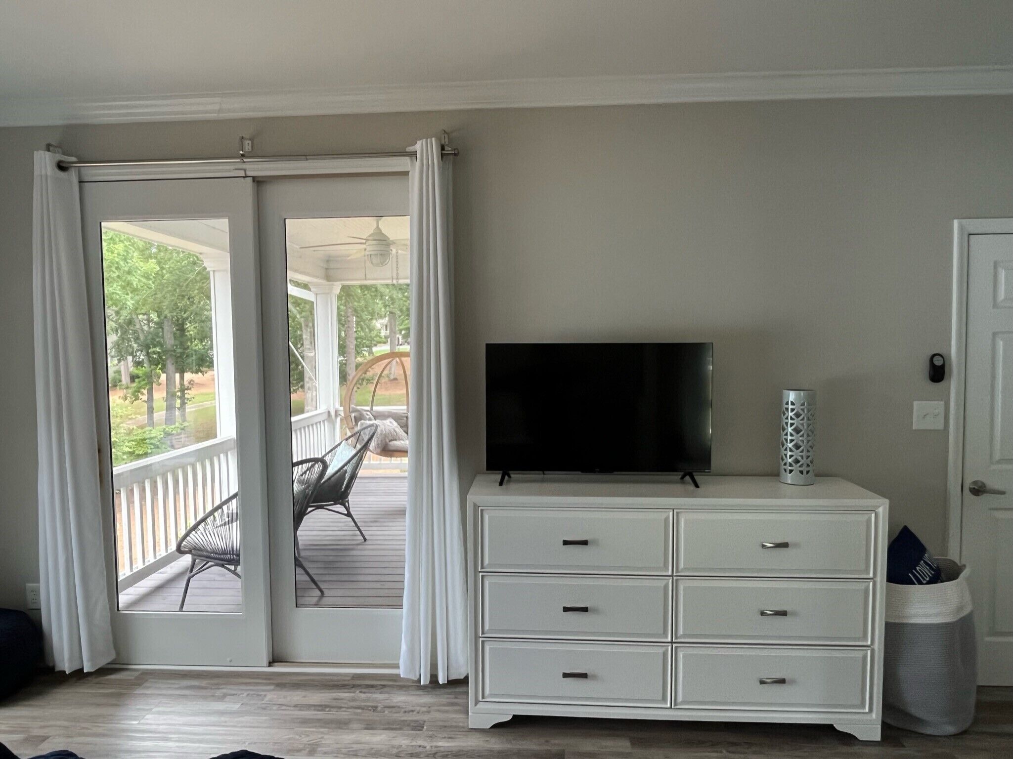 A room with a sliding glass door leading to an outdoor patio, a white dresser with drawers, a flat-screen TV on top, and a small decorative vase.