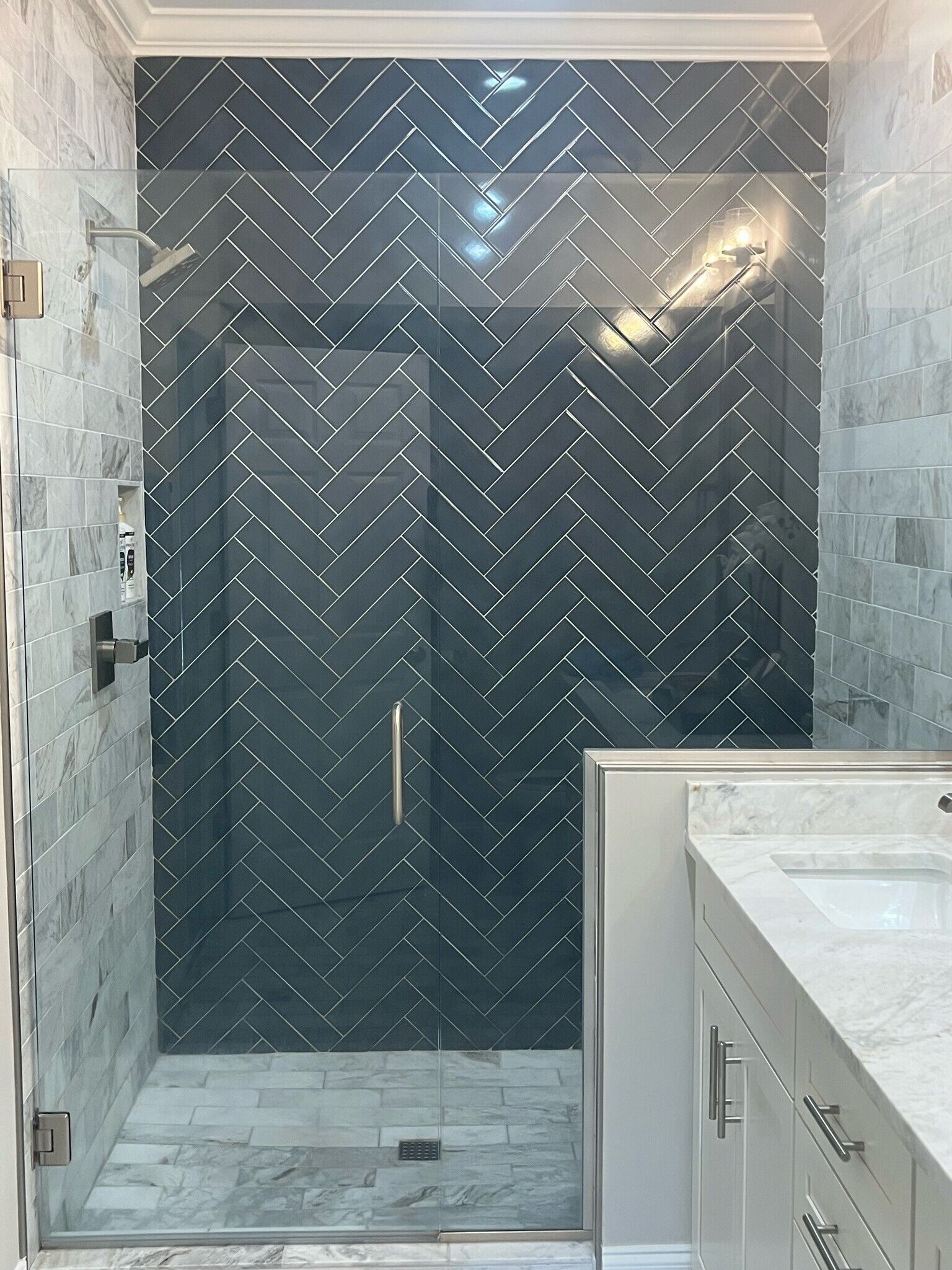 Une salle de bain moderne avec un carrelage disposé en motif chevron, une douche en verre et un lavabo surmonté d’un plan en marbre.
