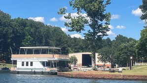 Una casa flotante amarrada en la orilla de un río, con una zona verde y árboles al fondo.