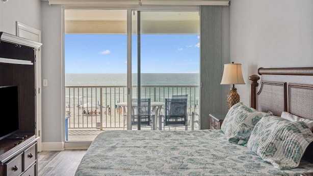 A bedroom with a large bed, a ceiling fan, a TV, and a view of the beach through a sliding door.