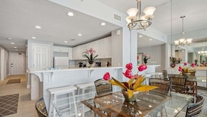 A modern kitchen with white cabinets, a glass dining table, wicker chairs, and a central island with pink flowers.