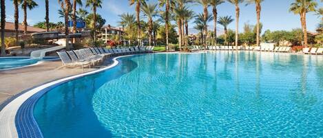A large, curved swimming pool with clear blue water, surrounded by palm trees and lounge chairs.
