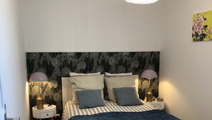 A bedroom with a bed, two bedside tables, a chair, a hanging light fixture, and a wall with a floral painting. - Cosy park near Grande Plage awarded 3-star label (Carnac)