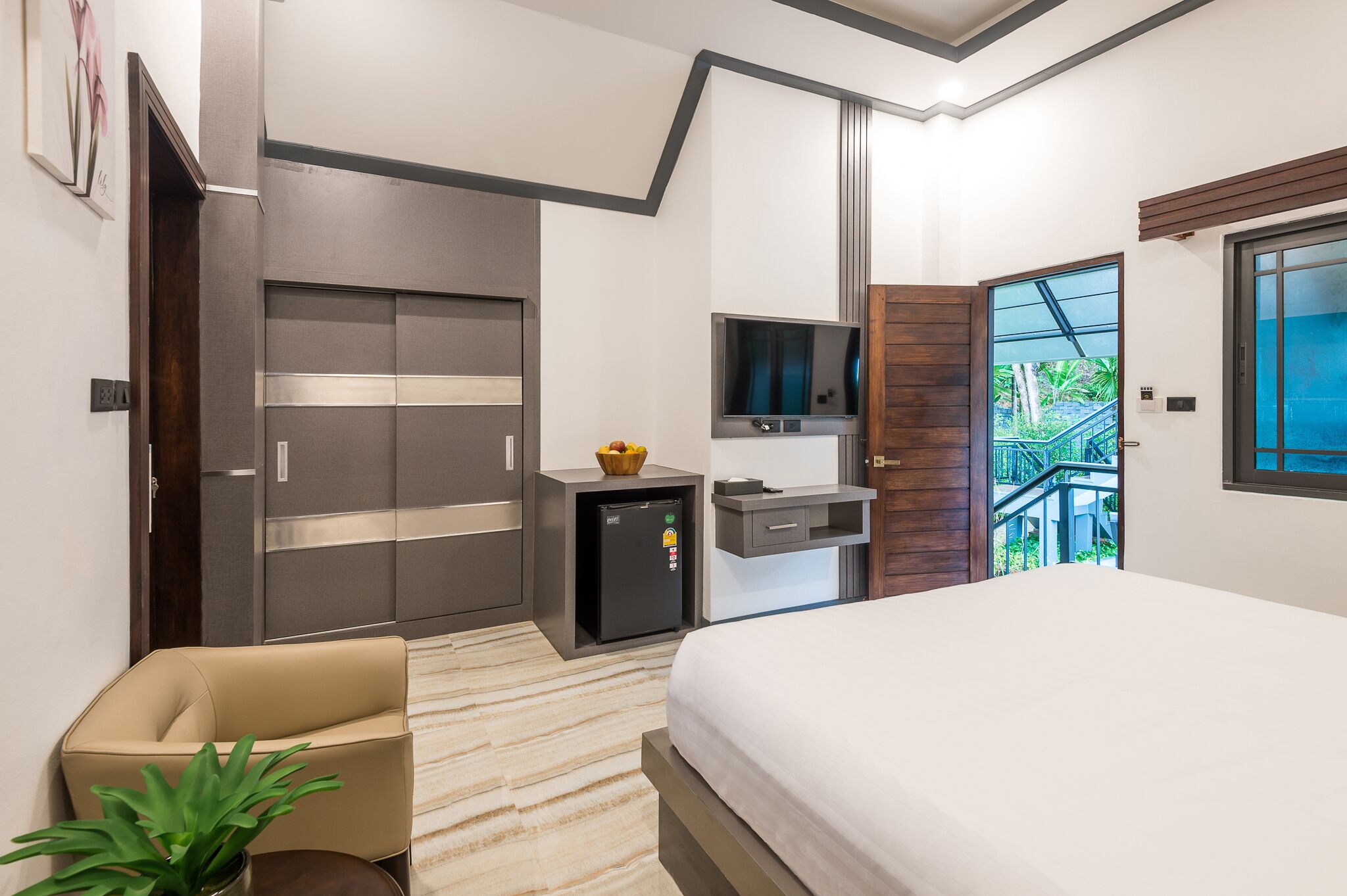 Une chambre d’hôtel moderne dotée d’un grand lit, d’un petit bureau avec une télévision, d’un minibar et d’un balcon offrant une vue sur la verdure.