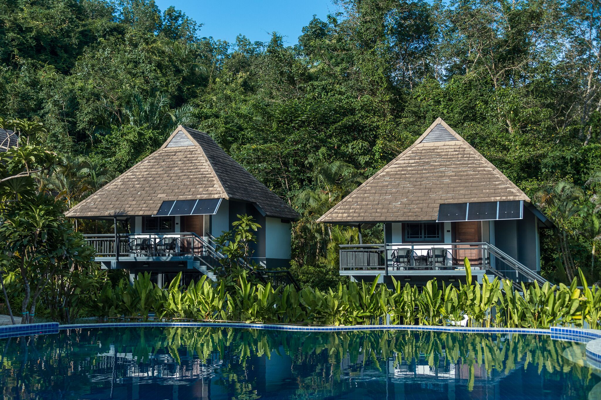 Blick auf einen Pool in einem Resort mit zwei bungalowartigen Gebäuden, jeweils mit einem Balkon und Solaranlagen.