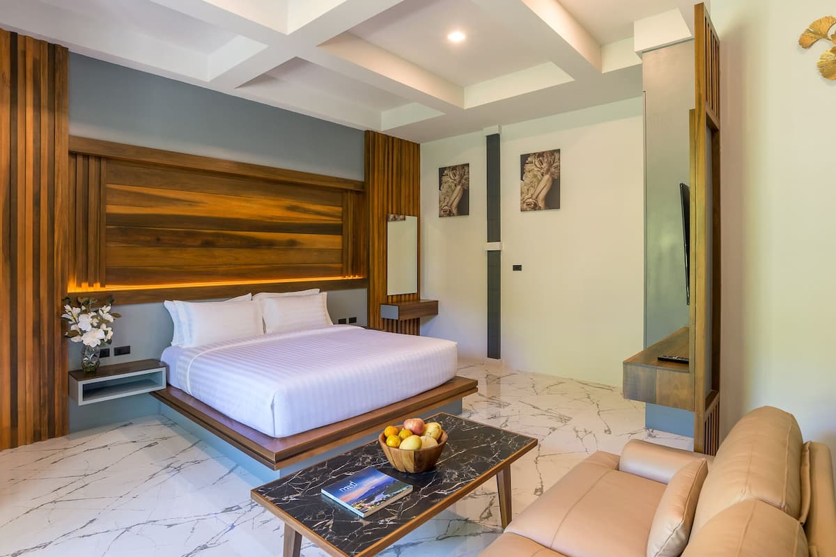 A modern hotel room with a large bed, a wooden headboard, a marble floor, a black coffee table with a bowl of fruit, and a beige sofa.