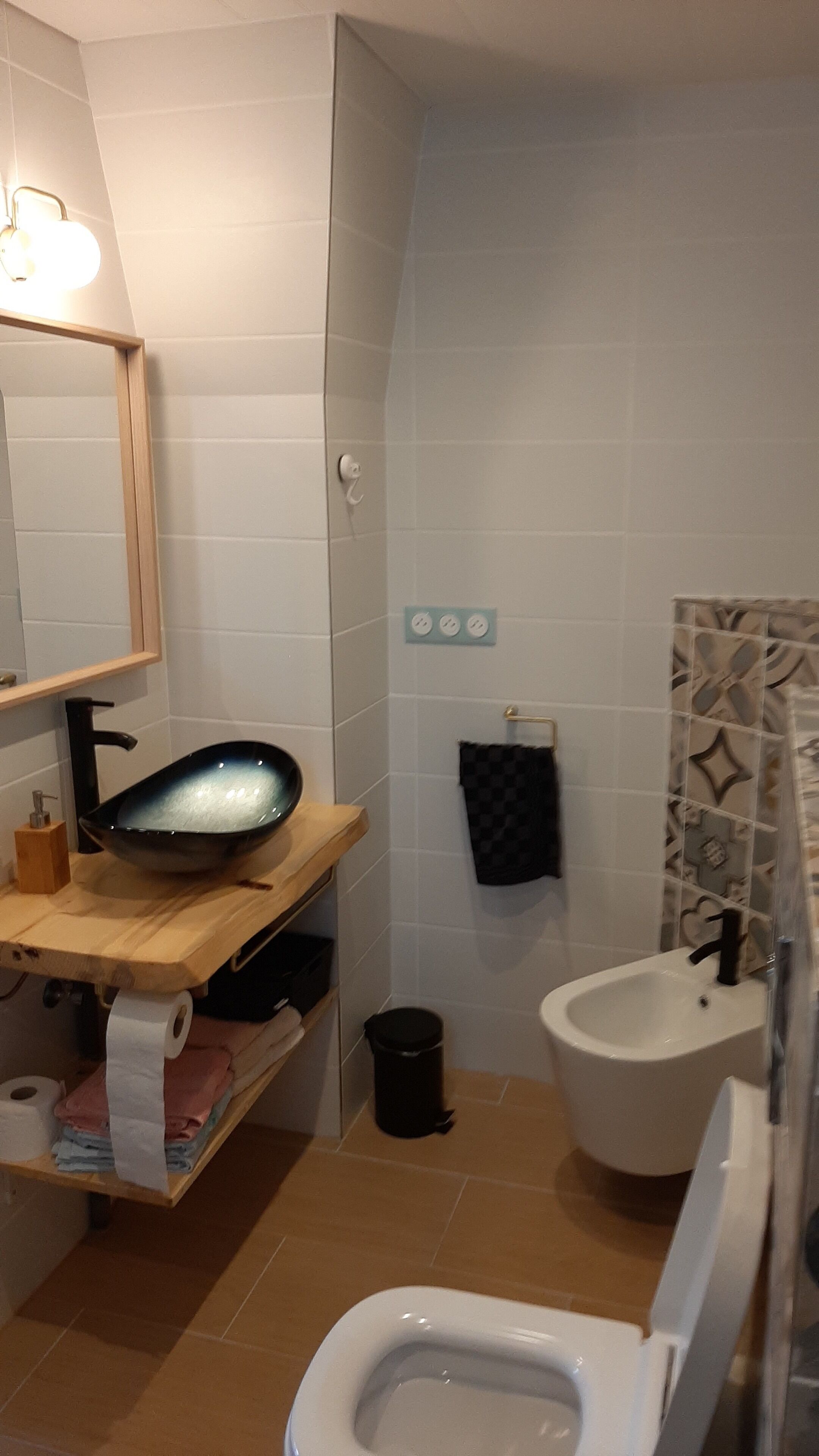 Une salle de bain équipée de toilettes, d’un bidet et d’un grand miroir. On y trouve une étagère avec des serviettes et une poubelle.