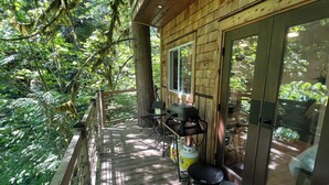 A wooden deck with a grill and a chair, surrounded by lush greenery. - Squamish Tree House (Brackendale)