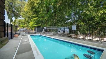Una piscina con un cartel que indica ausencia de socorrista, rodeada de árboles y un área vallada con sillas de descanso.
