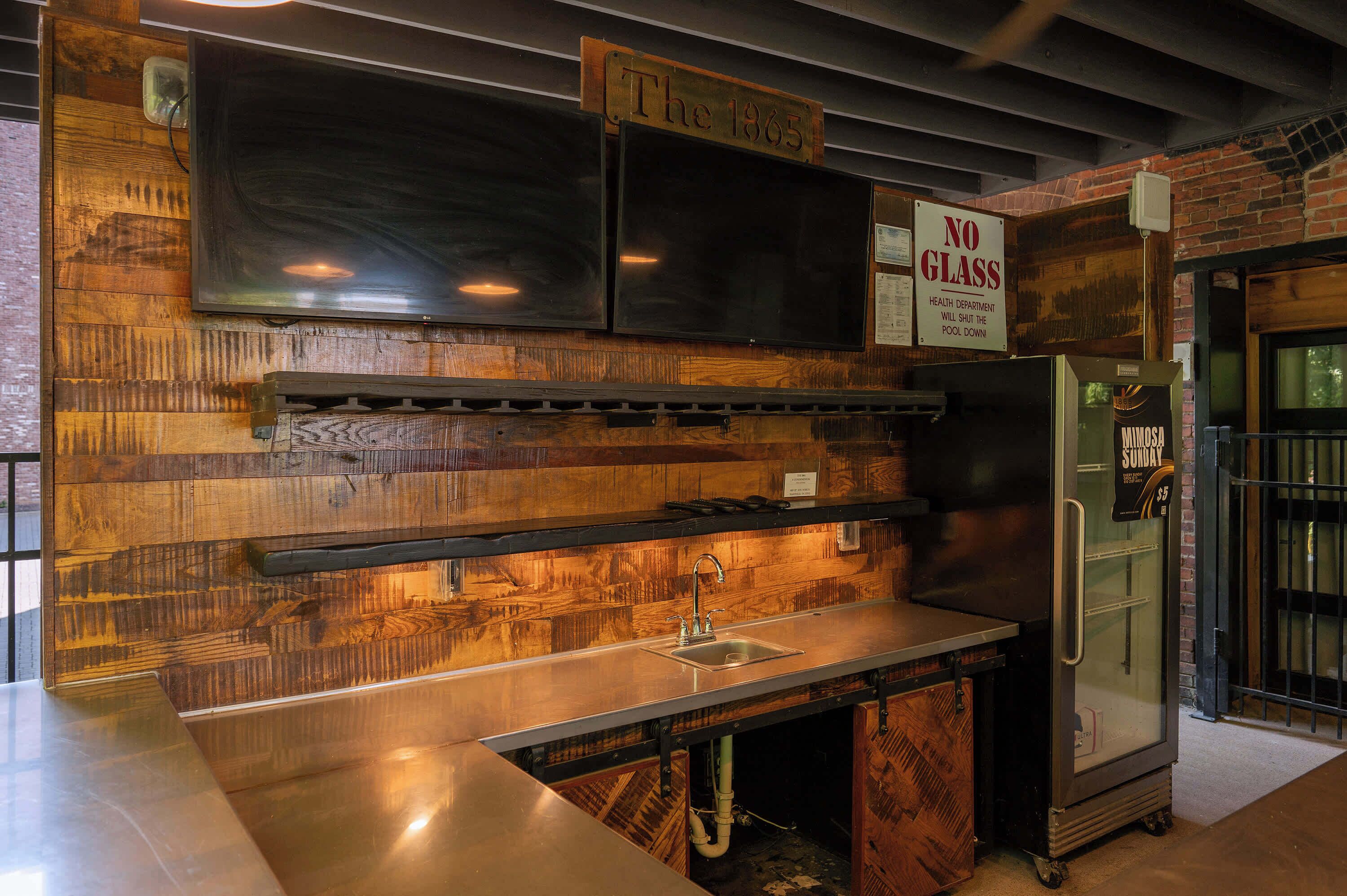 Una cocina con encimera de acero inoxidable, un lavamanos y un refrigerador. Hay un letrero que dice 'No Glass' y un letrero que dice 'The 1965' sobre el refrigerador.