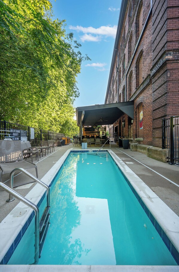 Una piscina con zona de estar cubierta, rodeada por un edificio de ladrillo y árboles.