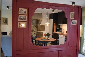 Ein Essbereich mit einem Holztisch, Stühlen und einer Vase mit Blumen. An der Wand hängen Bilder, und hinter dem Tisch ist eine Steinwand zu sehen.