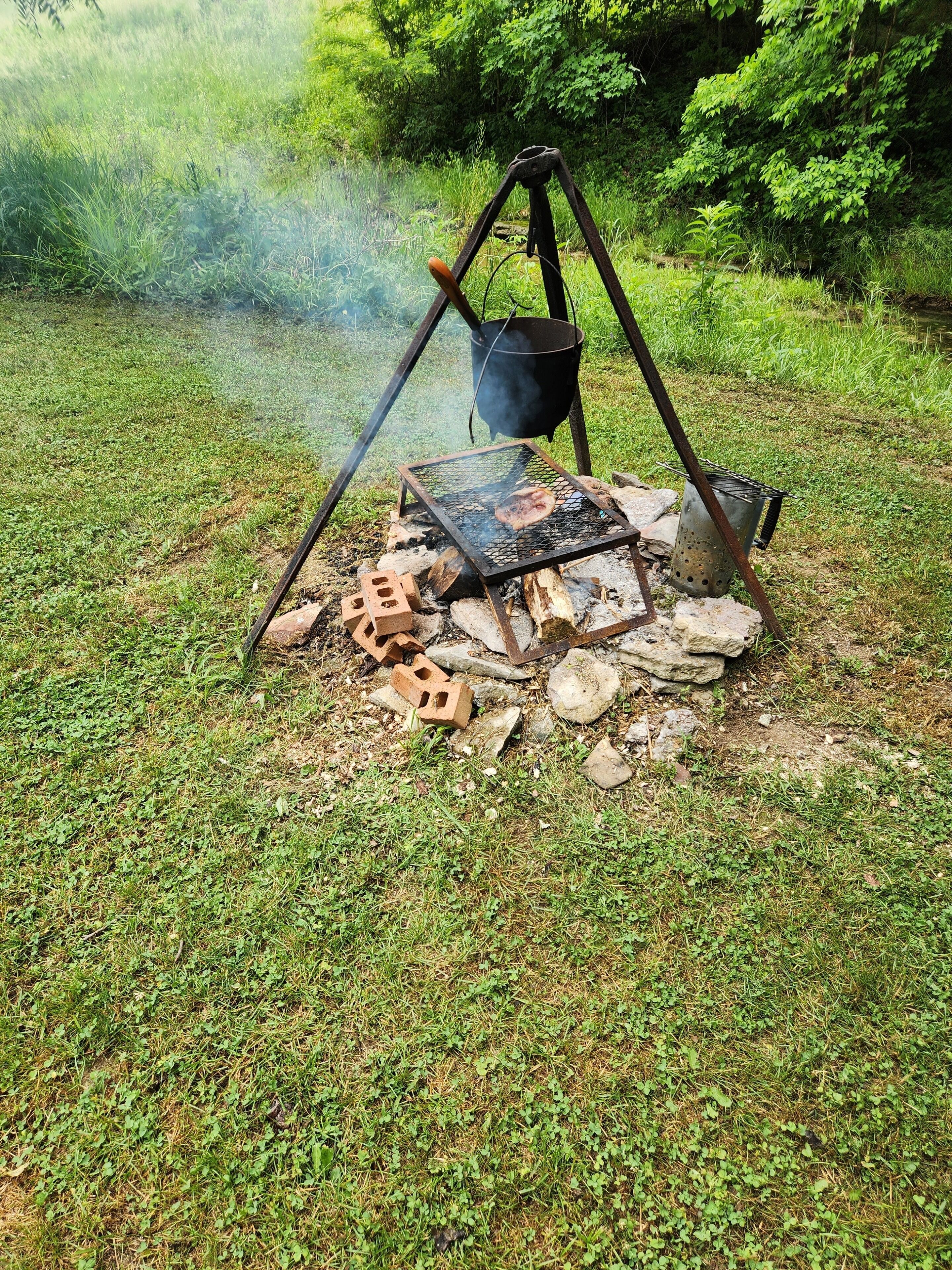 Een draagbare grill met een pan op een driepoot, omringd door stenen en bakstenen, gelegen in een grasrijke omgeving.
