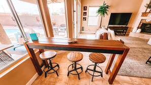 Un coin repas avec une table en bois et trois tabourets, un téléviseur et une cheminée, le tout dans une pièce bien éclairée grâce à de grandes fenêtres.
