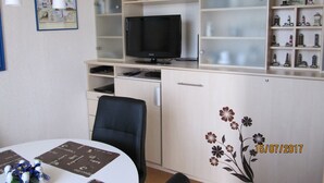 Ein Zimmer mit Schreibtisch, Stuhl, Fernseher und Regalen mit diversen Gegenständen.