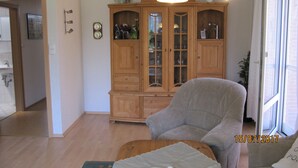 Ein Wohnzimmer mit einem Holzschrank, einem grauen Sessel und einem Tisch mit Kissen. Ein Fenster bietet Blick ins Freie.