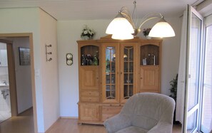 Un salon avec un meuble en bois, un fauteuil gris et une table avec un coussin. Il y a une fenêtre qui donne sur l’extérieur.