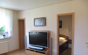 Ein kleines Zimmer mit Fernseher auf einem Schrank, einem Bett, einem Fenster mit Vorhängen und einem Bilderrahmen an der Wand.