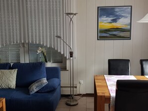 Uma sala de estar com um sofá azul, um abajur, uma mesa com cadeiras e um quadro na parede.