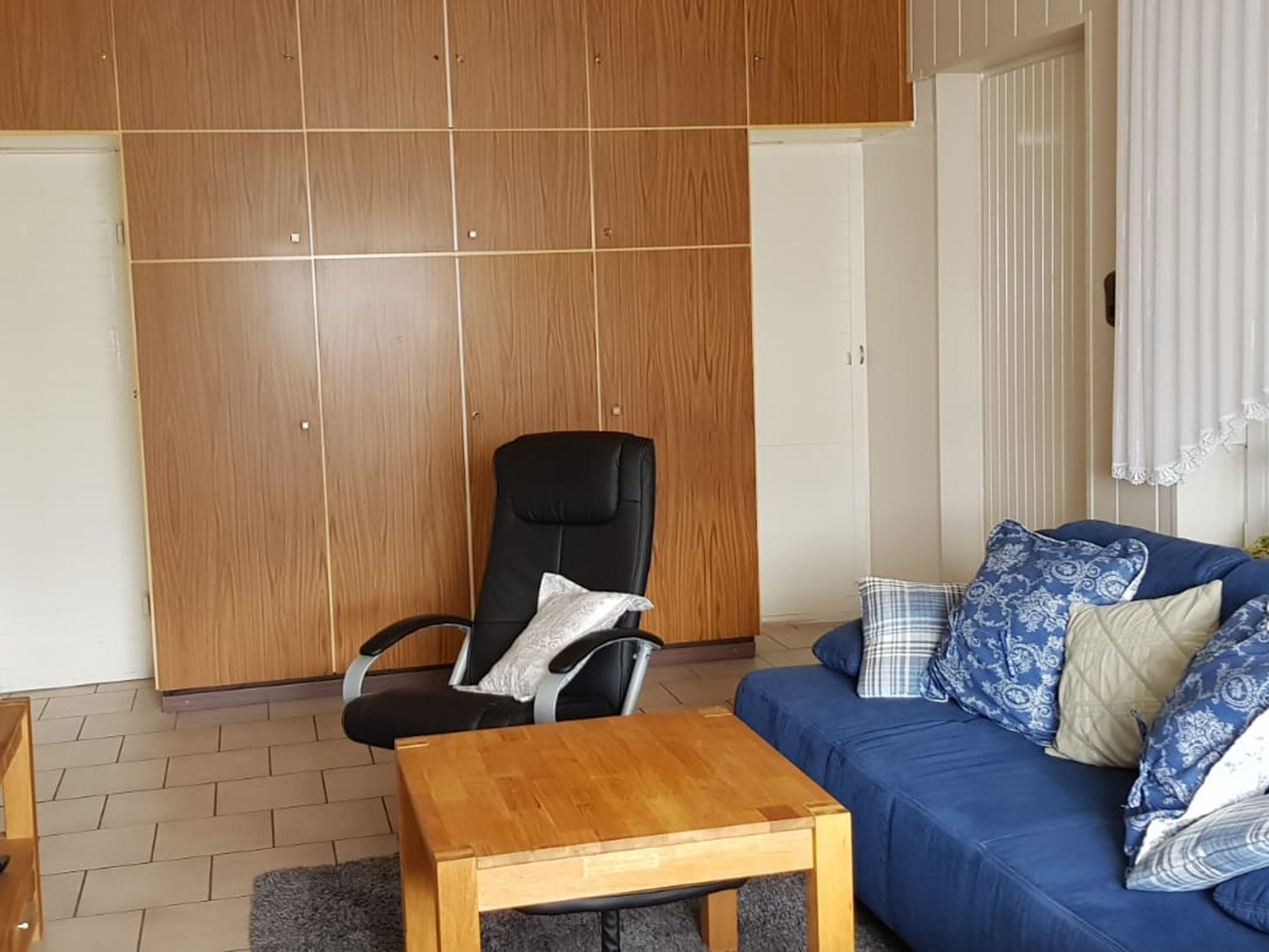 Une pièce comprenant une armoire en bois, un fauteuil de bureau noir, un canapé bleu avec des coussins à motifs et une table basse en bois.