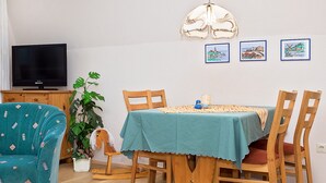 Ein Essbereich mit einem Tisch für vier Personen, umgeben von Holzstühlen. An der linken Wand ist ein Fernseher angebracht.