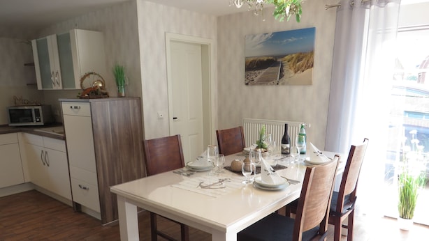 Een eetgedeelte met een witte tafel voor vier personen, houten stoelen, een keuken met kasten en een magnetron, en een schilderij aan de muur.