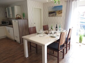 Un coin repas avec une table blanche dressée pour quatre personnes, des chaises en bois, un espace cuisine avec des placards et un four à micro-ondes, et un tableau accroché au mur.