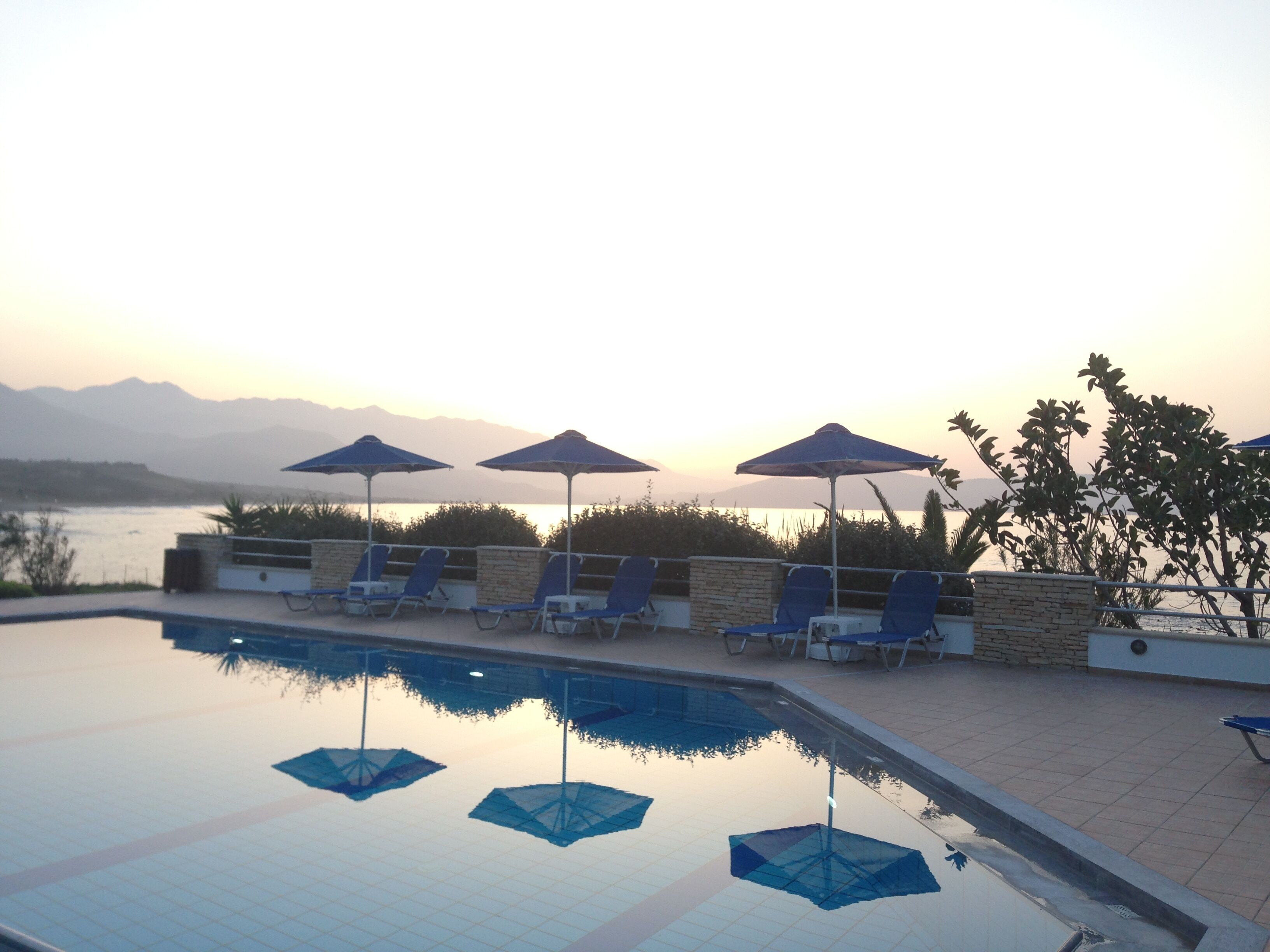 A poolside area with lounge chairs and umbrellas, overlooking a body of water and mountains.