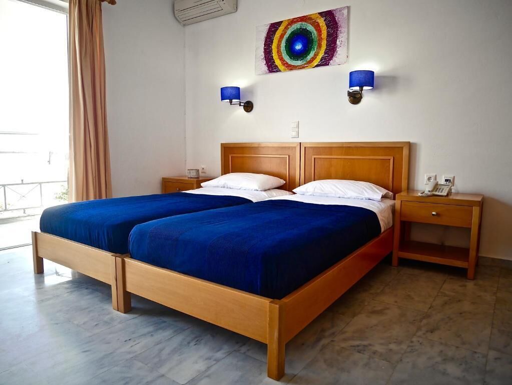 Een tweepersoonsbed met houten hoofdbord en voetenbord, twee nachtkastjes met lampen, een kleurrijk abstract schilderij aan de muur en een raam met gordijnen.