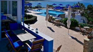 Un espace piscine aménagé sur le toit, avec des chaises et des parasols bleus, offrant une vue sur une mer d’un bleu limpide et sur des montagnes lointaines.