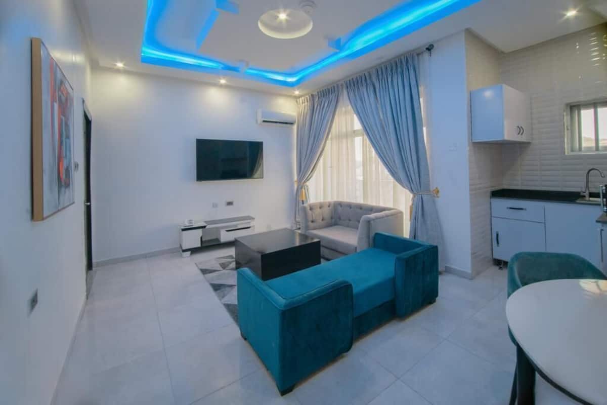 A modern living room with a sofa, a coffee table, and a TV. There is a kitchen area with cabinets and a sink.