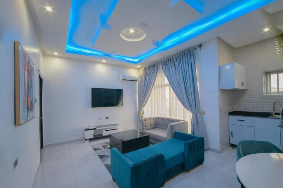 A modern living room with a sofa, coffee table, and TV. There is a kitchen area with cabinets and a window.