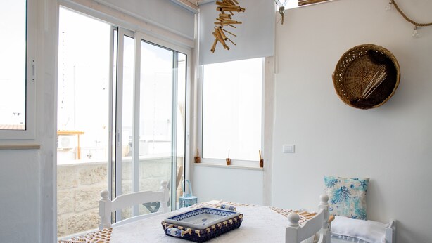 A room with a large window, a white bed with a blue and white tray, a white chair, and a small table with a blue basket.