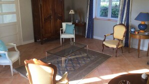 Uma sala de estar com uma mesa de centro de vidro, duas poltronas, um armário de madeira e uma janela com cortinas.