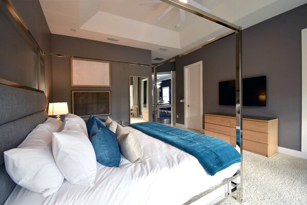 A modern bedroom with a large bed, a bedside table with a lamp, a dresser, and a television on the wall.