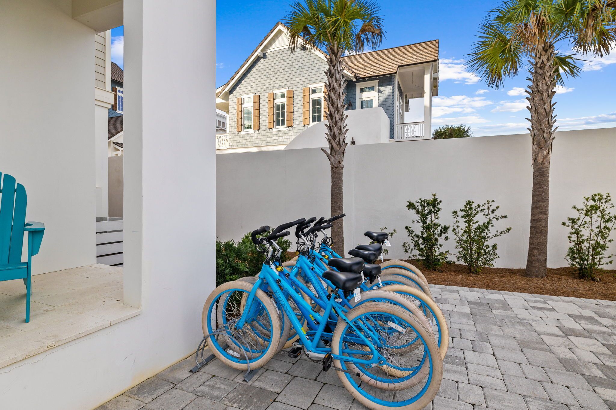 A blue bicycle parked in a covered outdoor area with a white wall and palm trees in the background.
