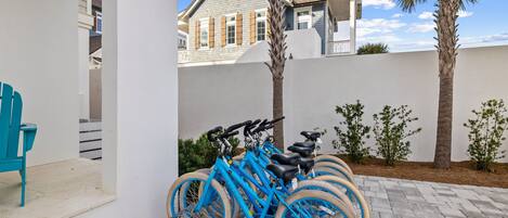 A blue bicycle parked in a covered outdoor area with a white wall and palm trees in the background.