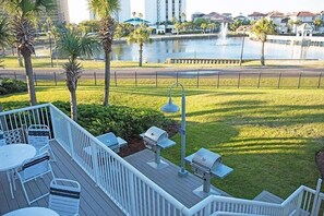 A deck with outdoor seating and barbecue grills, overlooking a landscaped area with palm trees and a body of water. - Terrace at Pelican Beach 1005 (2-Bedroom Condo) with Pool by Pelican Beach Management (Destin)