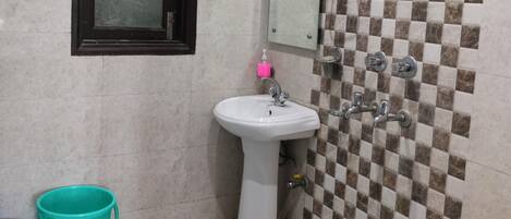 A bathroom with a pedestal sink, a mirror, a shower with a checkered tile wall, a fan, and a blue bucket.
