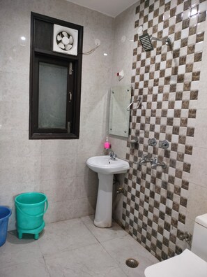 Un baño con lavamanos pedestal, espejo, ducha con pared de baldosas a cuadros, un ventilador y un cubo azul.