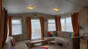 A cozy room with a patterned sofa, a small table, and a window with curtains. - A&P Sheron Holiday Home - Millfields (Skegness)