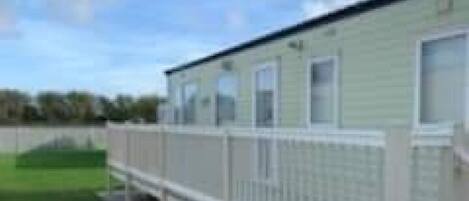 A row of single-story mobile homes with white railings, set against a backdrop of a grassy field and a clear sky.